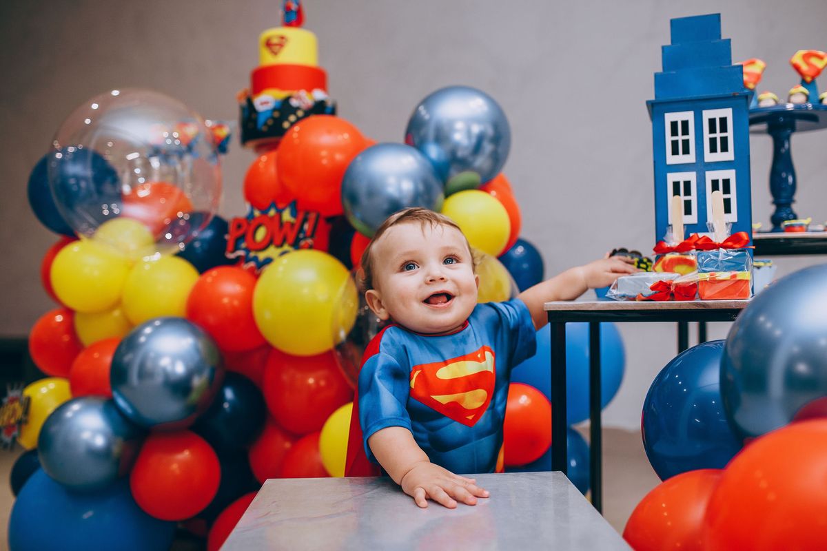 Festa infantil com tema de super-herói: bebê fantasiado diante da mesa decorada com balões